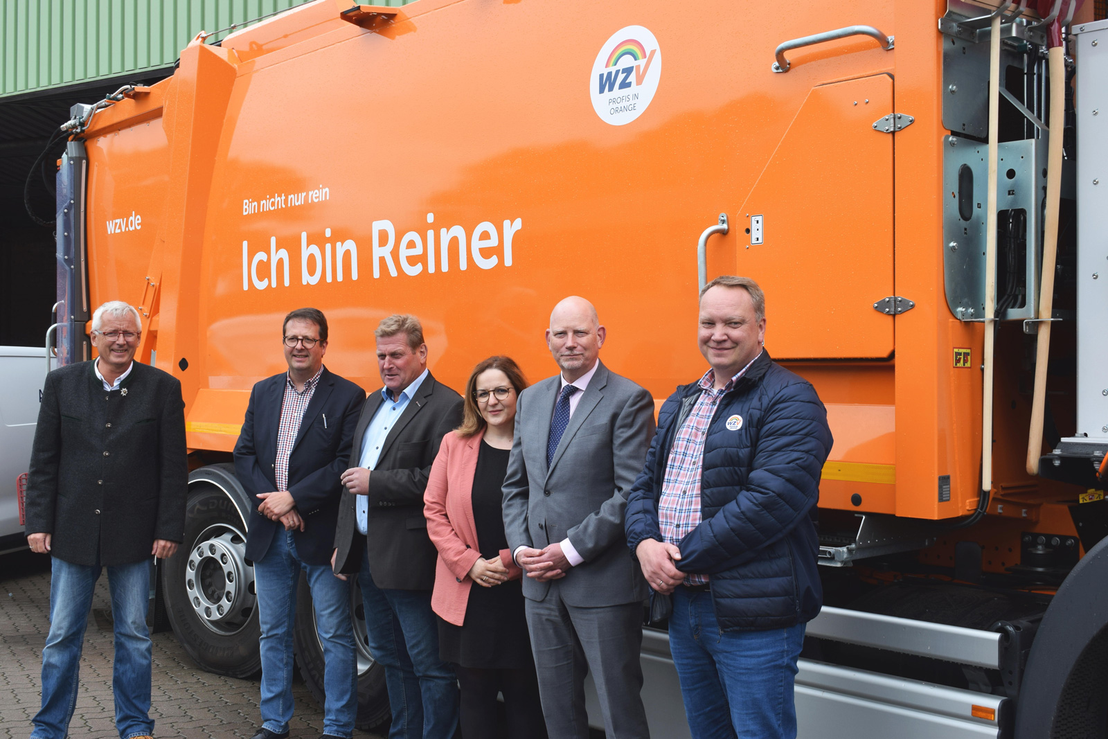 Sechs Personen stehen nebeneinander vor einem orangefarbenen Müllwagen mit der Aufschrift 'Ich bin Reiner' und dem Logo von WZV