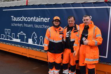 Drei Personen in orangefarbener Arbeitskleidung stehen vor einem großen blauen Container mit der Aufschrift 'Tauschcontainer - schenken, freuen, tauschen